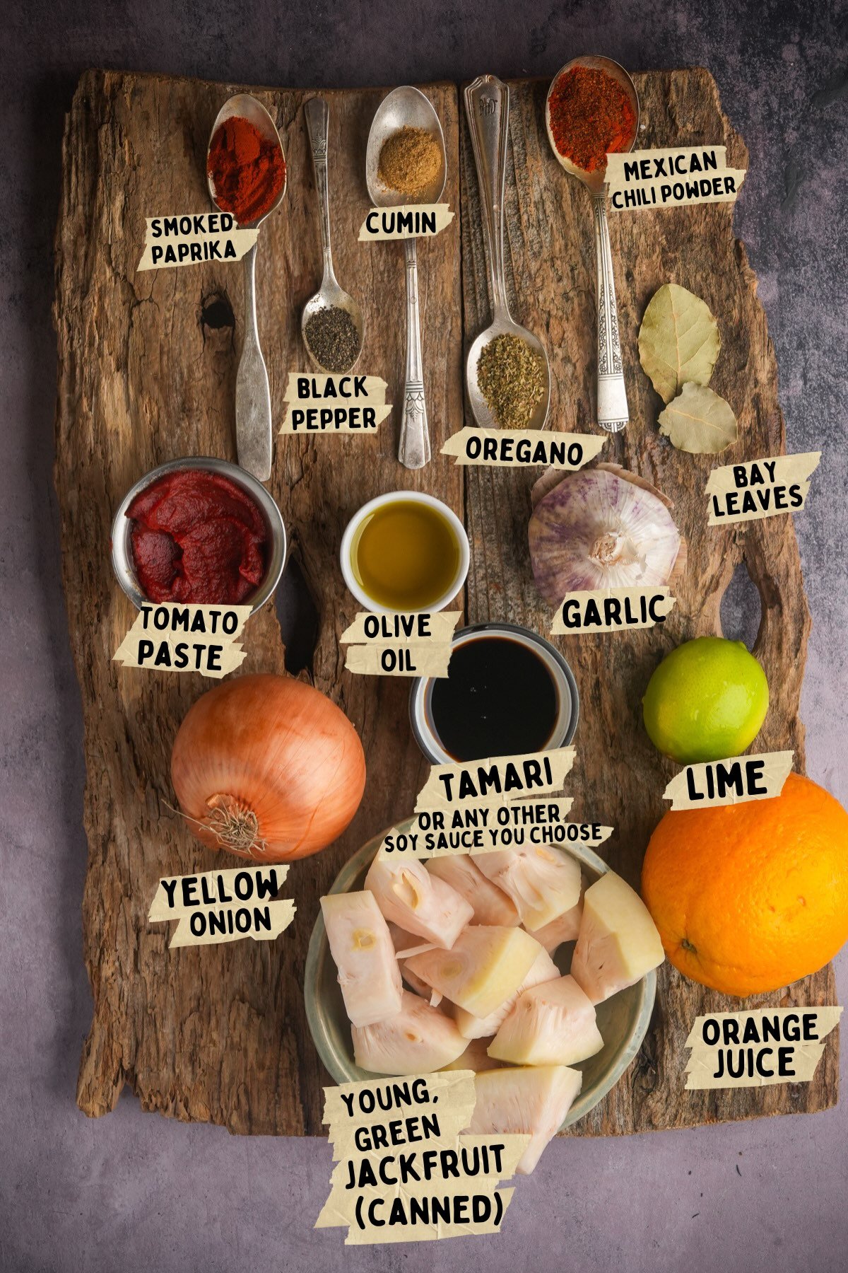 A wooden board with various ingredients labeled for a vegan carnitas recipe: smoked paprika, cumin, black pepper, oregano, Mexican chili powder, bay leaves, tomato paste, olive oil, garlic, onion, tamari, lime, orange juice, and canned jackfruit.