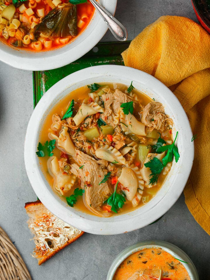 A white bowl filled with vegetable and shredded chicken soup, garnished with fresh herbs, sits on a table alongside other bowls of soup and a piece of toasted bread.