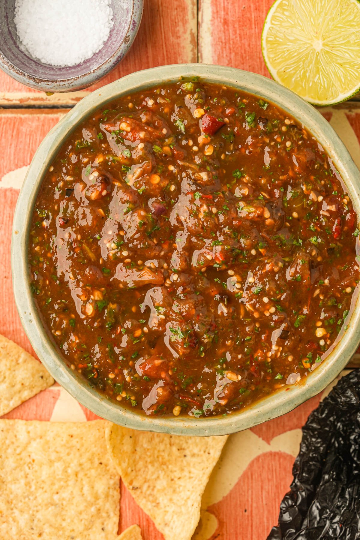 A bowl of chunky red chile de arbol salsa sits on a tiled surface, surrounded by tortilla chips, a halved lime, a dish of salt, and a dried chili pepper.