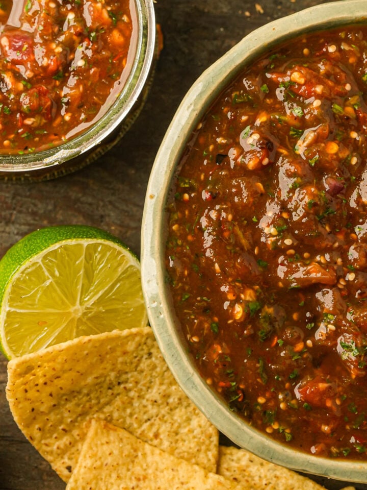 A bowl and jar of chunky chile de arbol salsa with tortilla chips and a halved lime on a wooden surface.