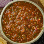 A bowl of chunky red chile de arbol salsa with visible herbs and seeds, surrounded by tortilla chips and a partial view of sliced red onion.