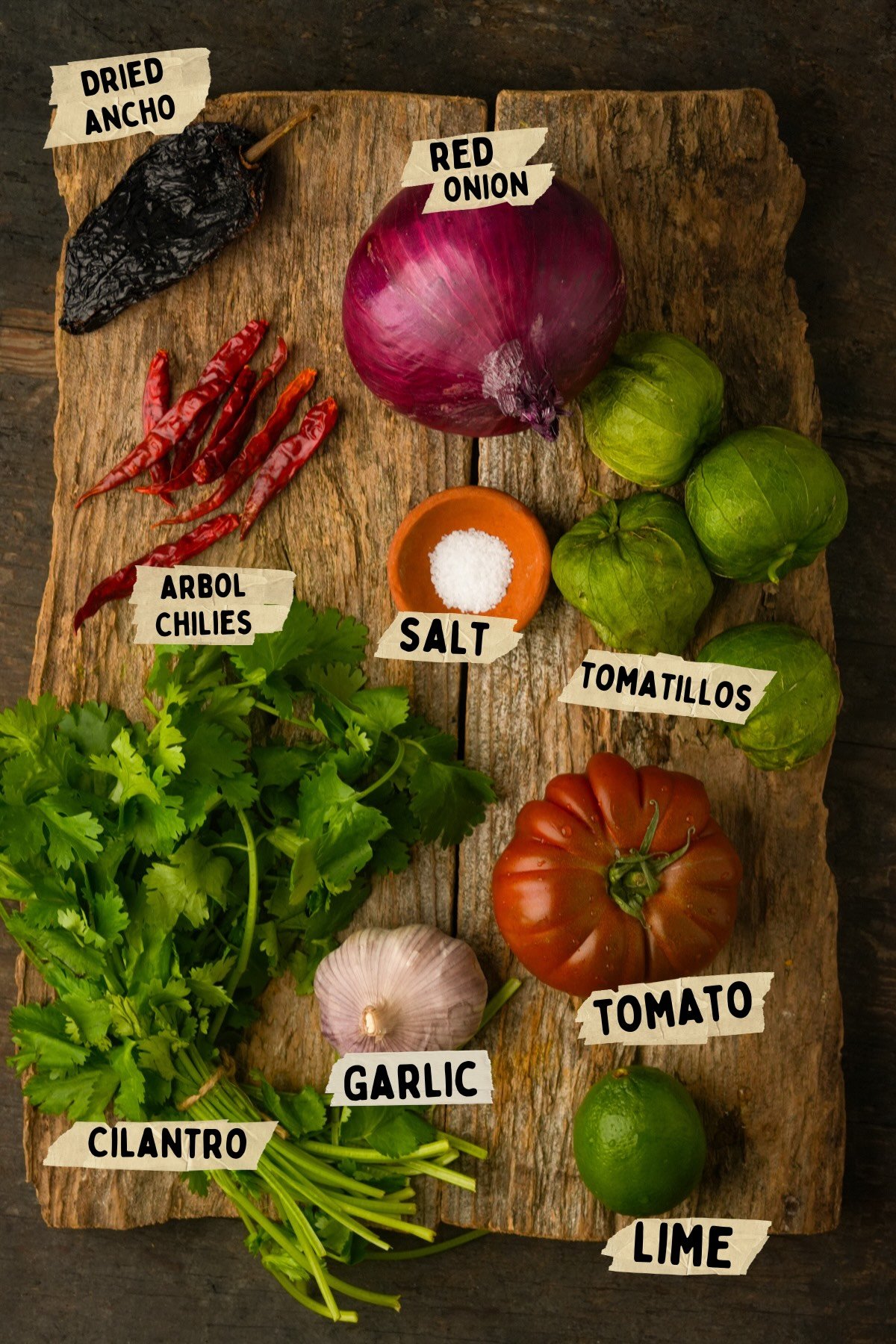 A wooden board displaying chile de arbol salsa recipe ingredients: dried ancho, arbol chiles, red onion, salt, tomatillos, cilantro, garlic, tomato, and lime, each labeled with handwritten tags.