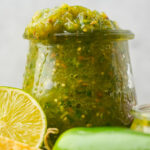 A glass jar filled with green jalapeño salsa sits beside a halved lime and green chili peppers.