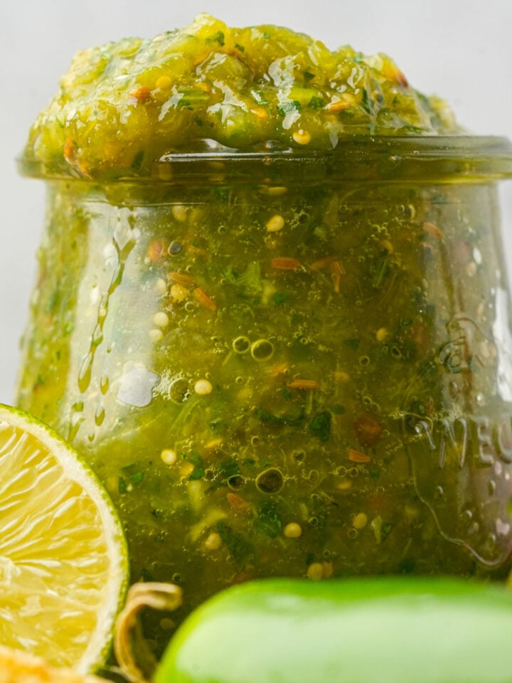 A glass jar filled with green jalapeño salsa sits beside a halved lime and green chili peppers.