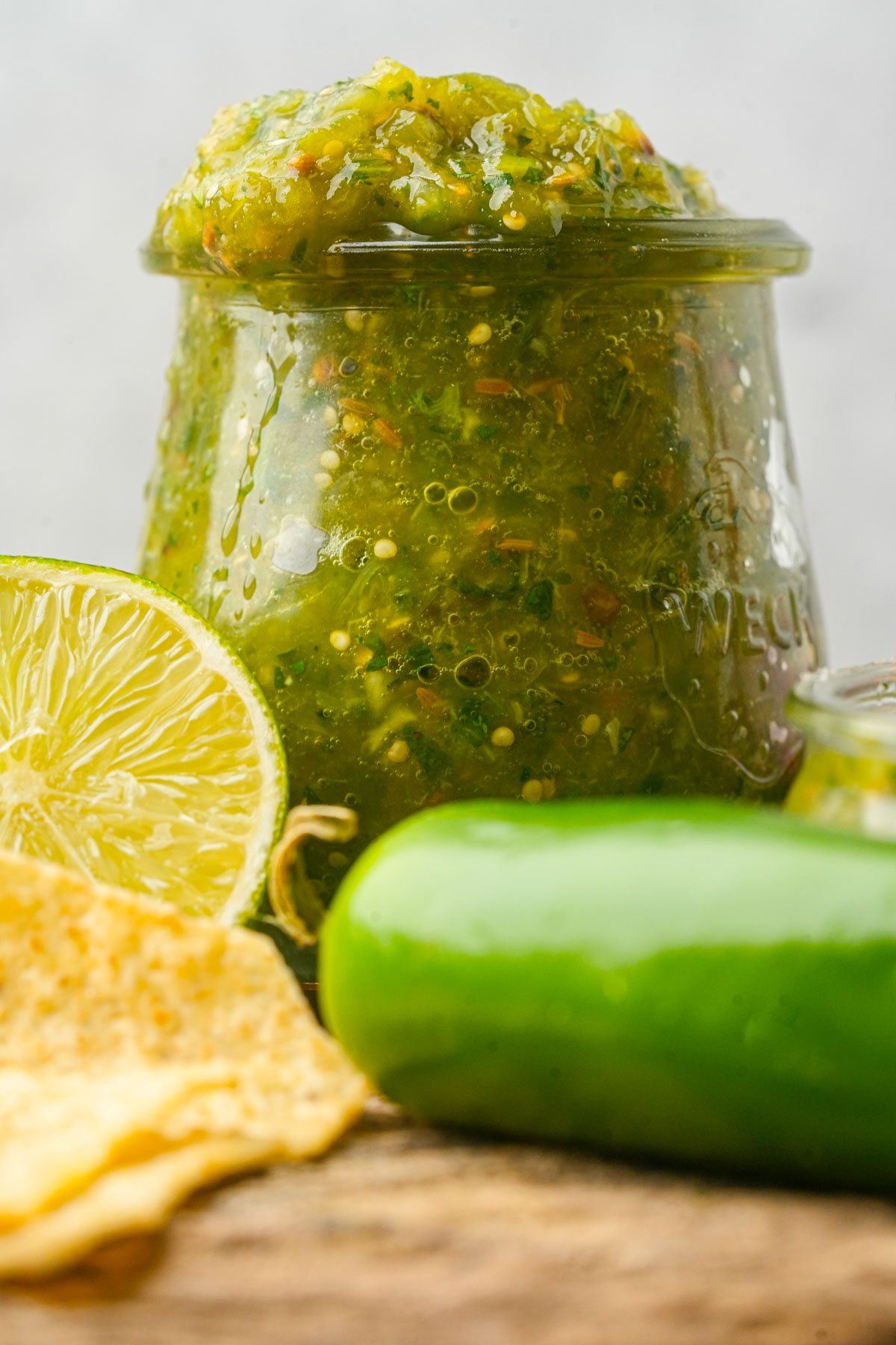 A jar of green jalapeño salsa sits next to a sliced lime, a jalapeño pepper, and a tortilla chip on a wooden surface.
