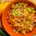 A bowl of avocado mango salsa with tomatoes, red onion, cilantro, and jalapeño, in an orange dish on a wooden surface.