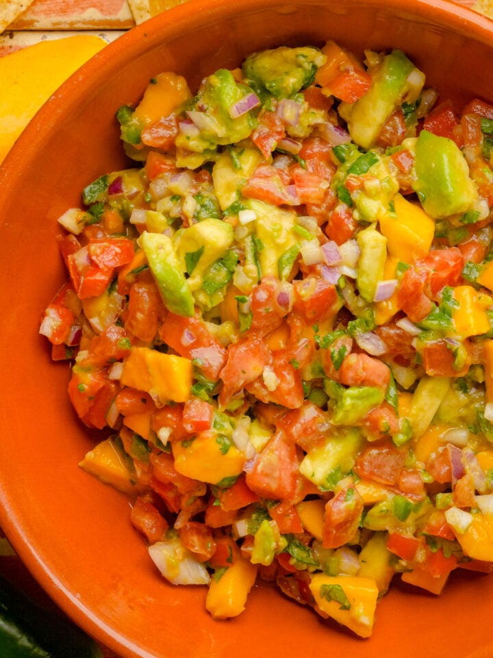 A bowl of avocado mango salsa with tomatoes, red onion, cilantro, and jalapeño, in an orange dish on a wooden surface.