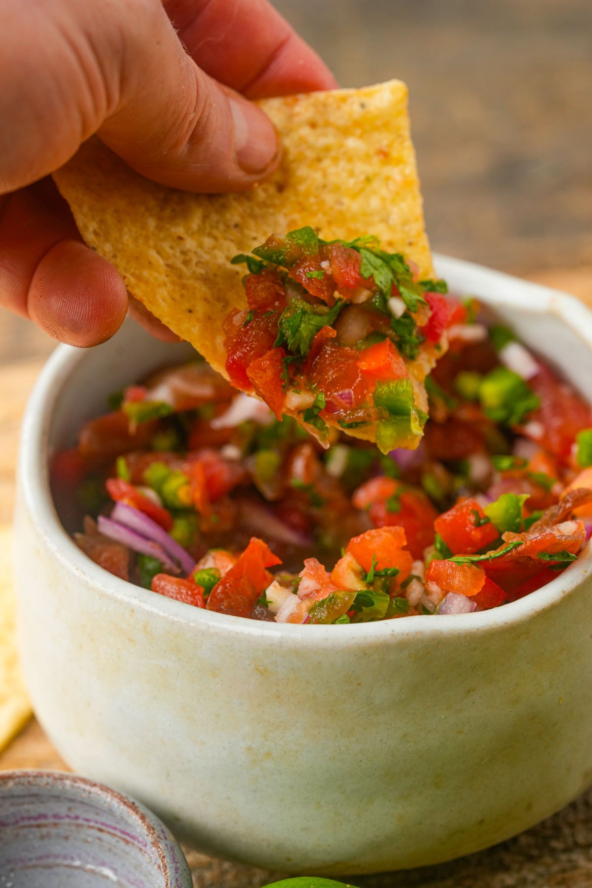 A hand dips a tortilla chip into a bowl of fresh salsa fresca made with tomatoes, onions, cilantro, and jalapeños.