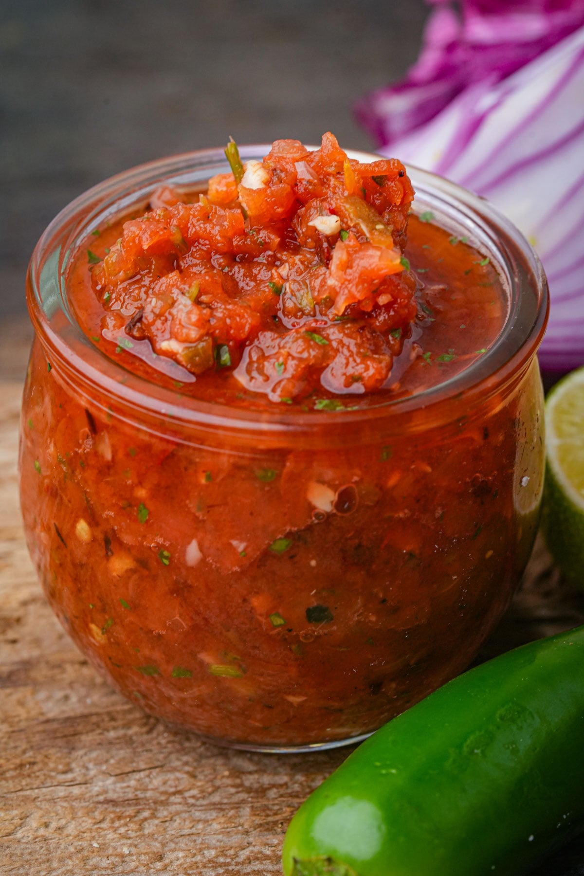 A glass jar filled with chunky salsa ranchera sits on a wooden surface, with a whole jalapeño, a lime wedge, and purple cabbage in the background.