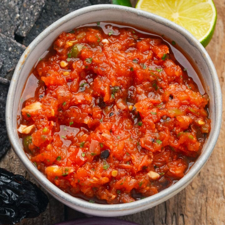 A bowl of chunky salsa ranchera with visible herbs and diced vegetables, placed on a wooden surface next to a sliced lime and blue corn tortilla chips.