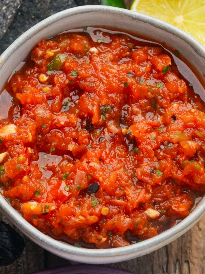 A bowl of chunky salsa ranchera with visible herbs and diced vegetables, placed on a wooden surface next to a sliced lime and blue corn tortilla chips.