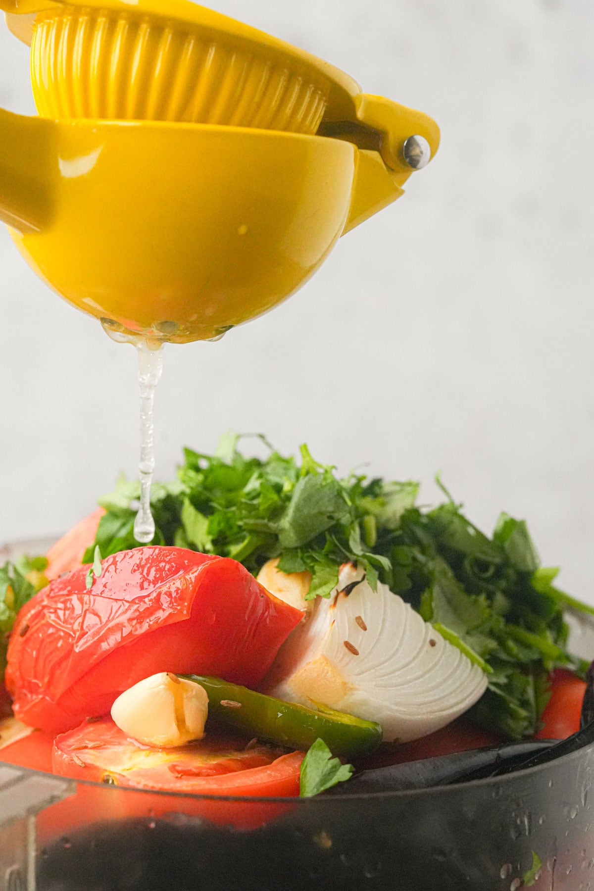 A yellow citrus squeezer drips lime juice onto chopped tomatoes, onion, garlic, green chili, and fresh herbs in a food processor.