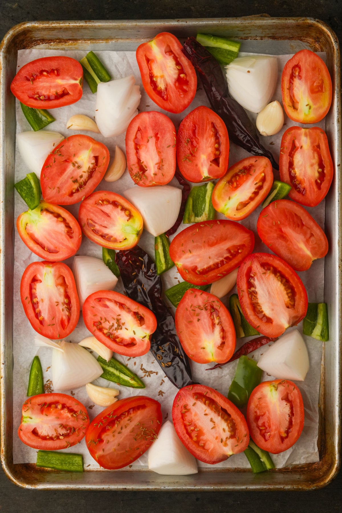A baking sheet with halved Roma tomatoes, chopped white onion, garlic cloves, dried chili peppers, and green bell pepper pieces arranged on parchment paper.