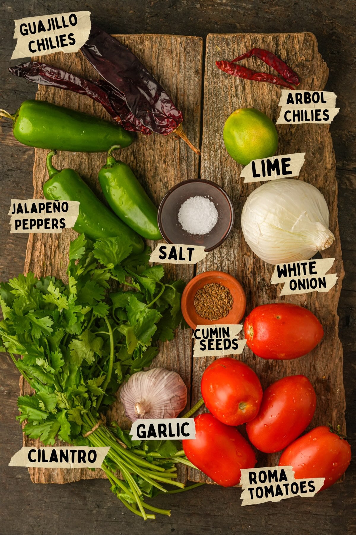 A wooden board displays ingredients for a salsa ranchera recipe: guajillo and arbol chiles, jalapeno peppers, lime, salt, white onion, cilantro, garlic, cumin seeds, and roma tomatoes with labeled tags.