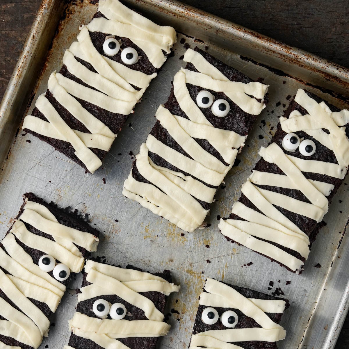 Six rectangular brownies decorated with strips of white frosting and candy eyes to resemble mummies, arranged on a metal baking sheet.