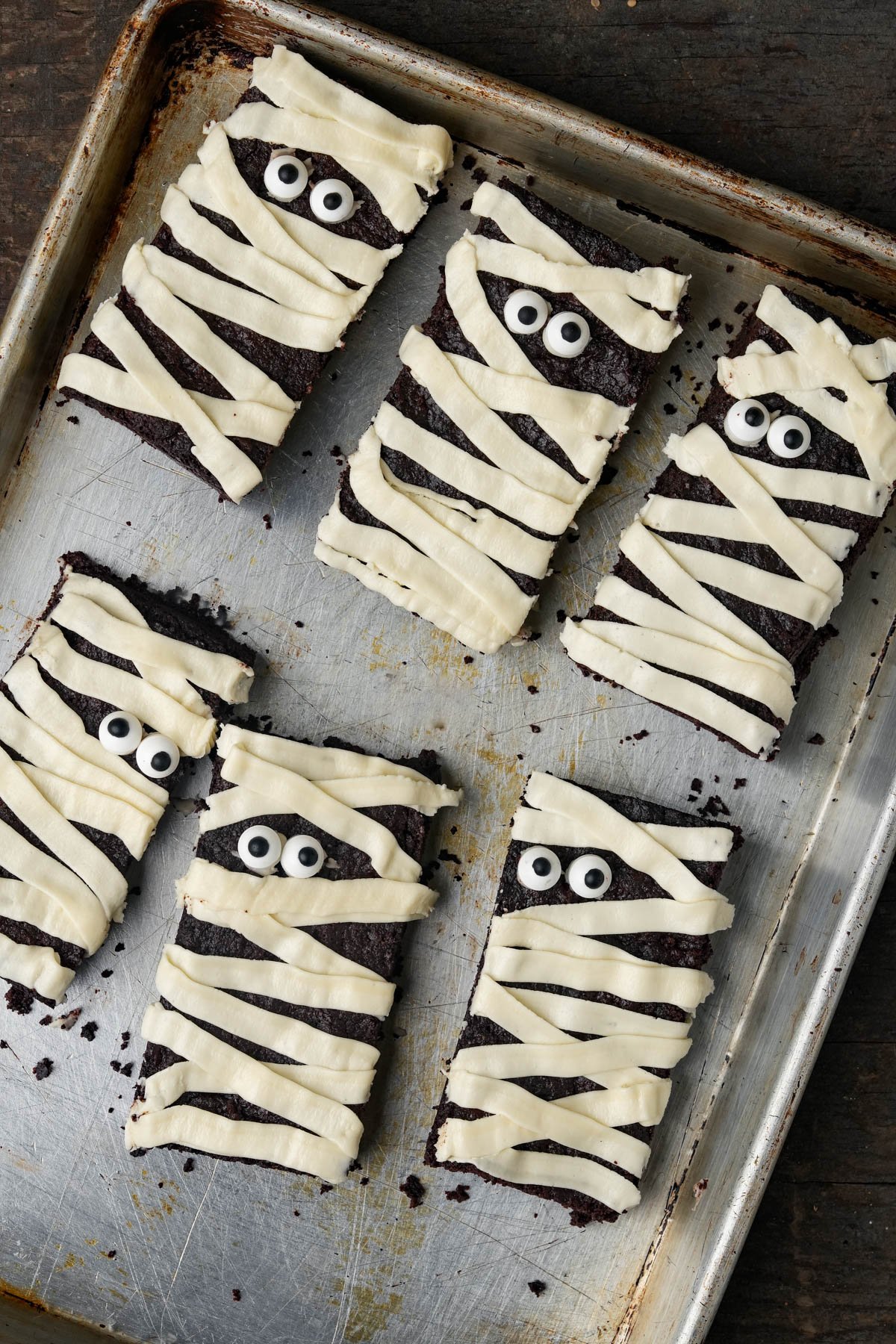 Six rectangular brownies decorated with white icing to resemble mummies, featuring candy eyes, arranged on a metal baking sheet.