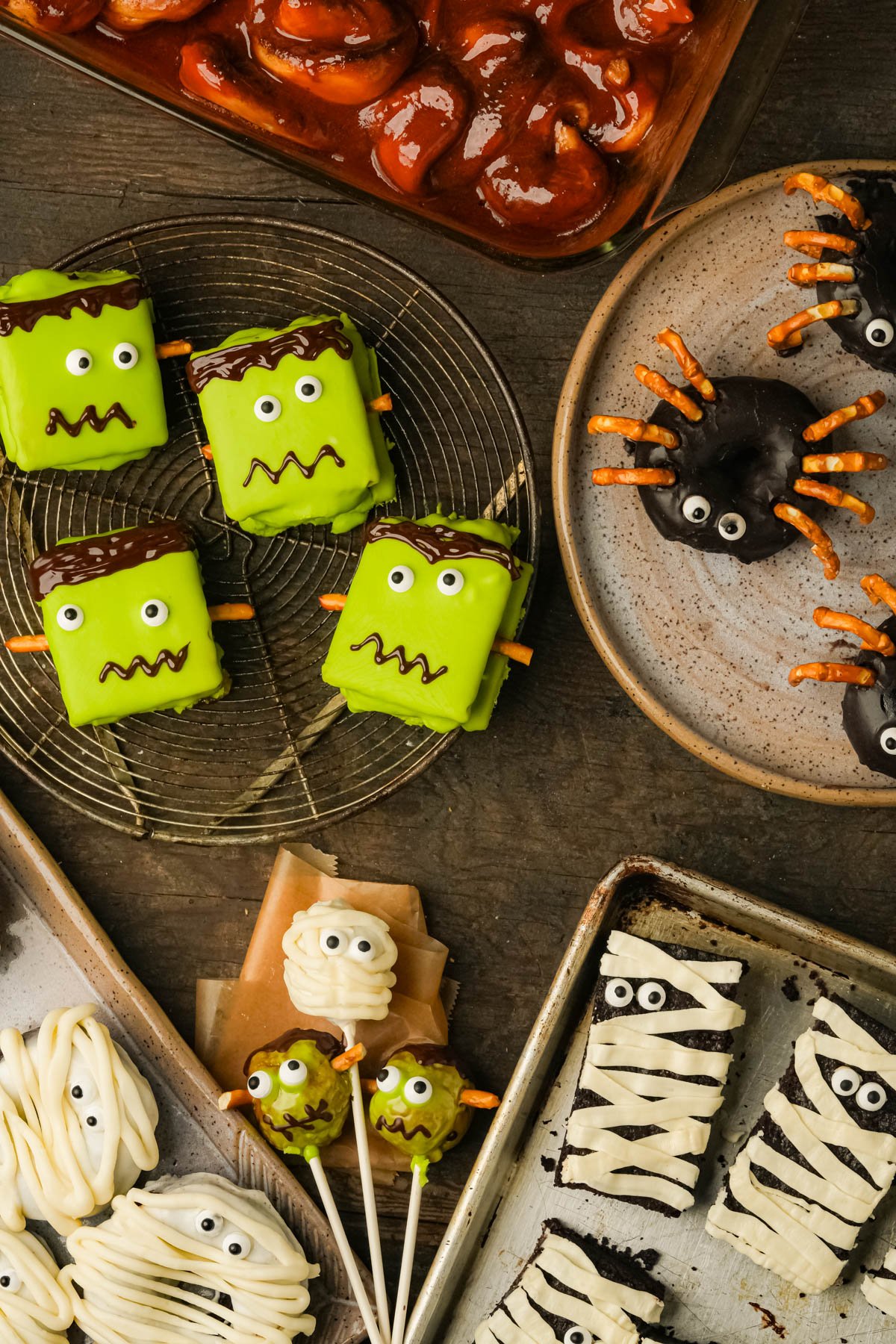 Assorted Halloween-themed treats including green Frankenstein s'mores, spider donuts with pretzel legs, mummy cake pops, and a dish of cinnamon roll guts.