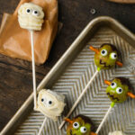 Halloween cake pops decorated as a white mummy and green monsters with candy eyes and pretzel stick ears, arranged on a baking sheet and parchment paper.