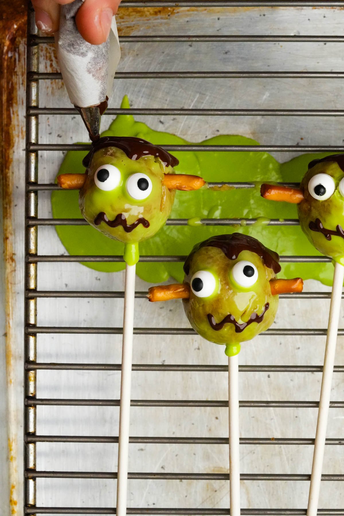 Hand decorating green Vegan Halloween Cake Pops with candy eyes, pretzel stick ears, and chocolate drizzle on a wire rack over a baking sheet.