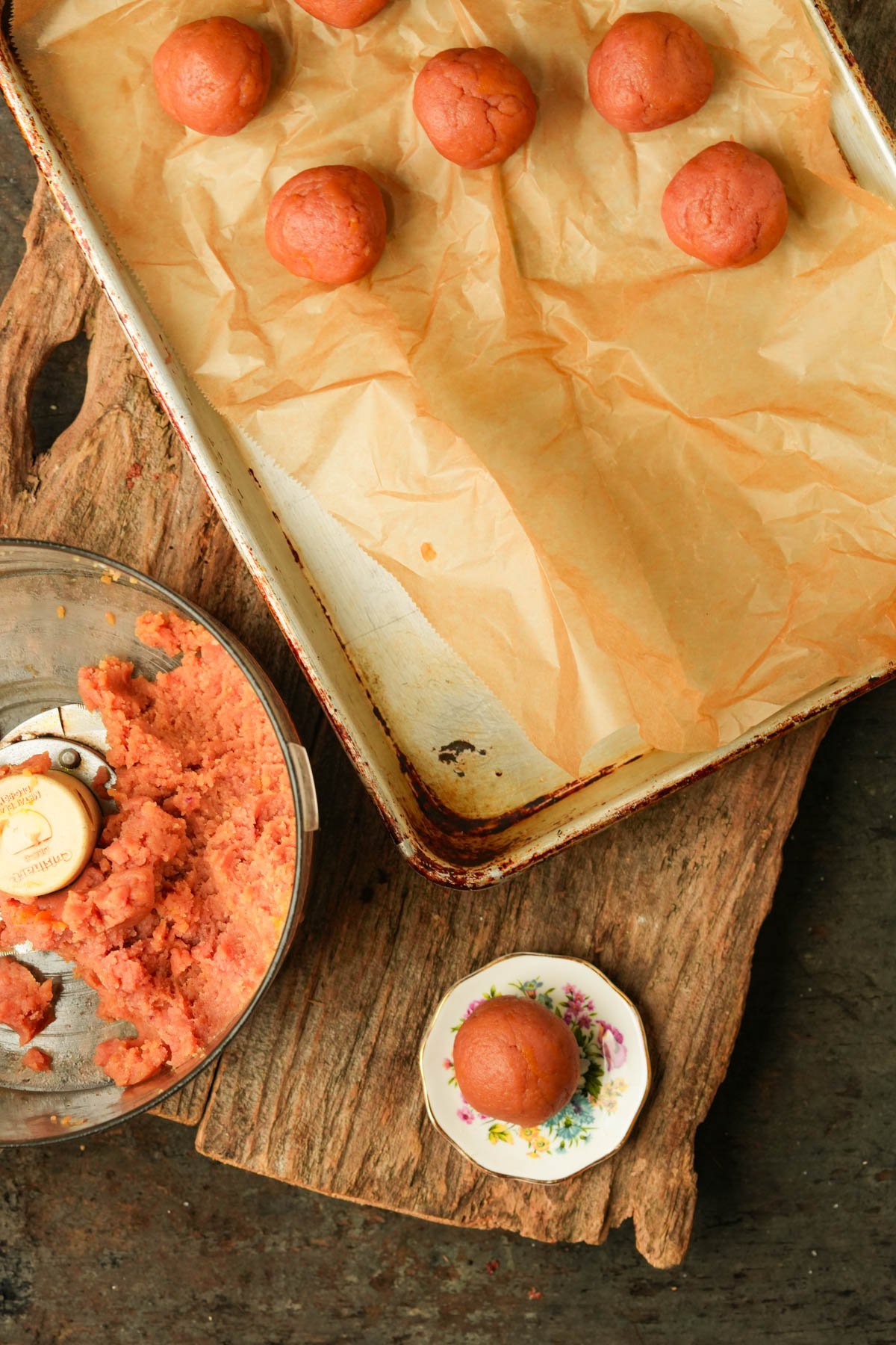 A baking tray with parchment paper holds several round dough balls. Nearby are a food processor with pink dough and a small floral plate with another dough ball.