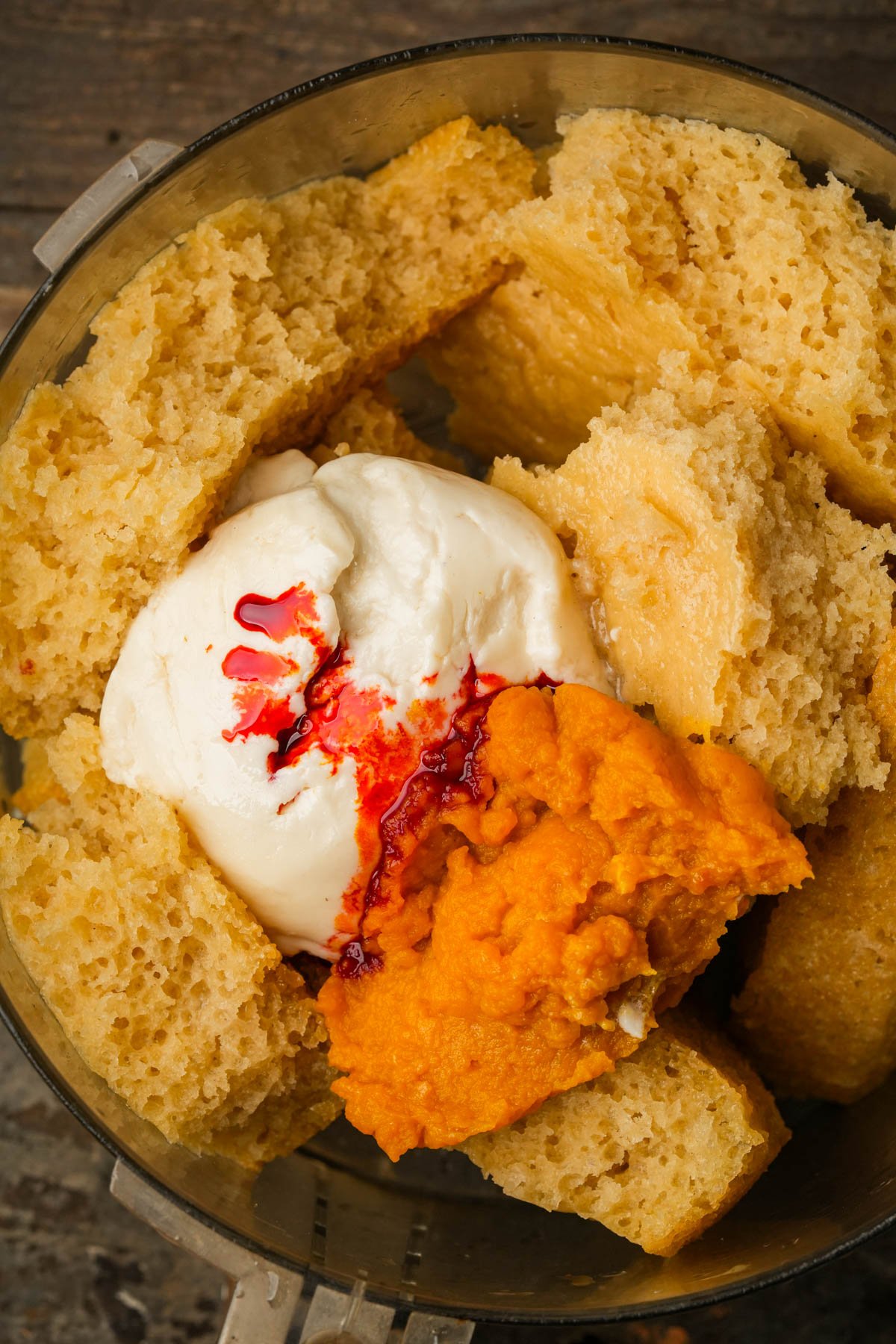 Chunks of sponge cake, a dollop of vegan cream cheese, pumpkin puree, and red food coloring are placed in a food processor bowl on a wooden surface.