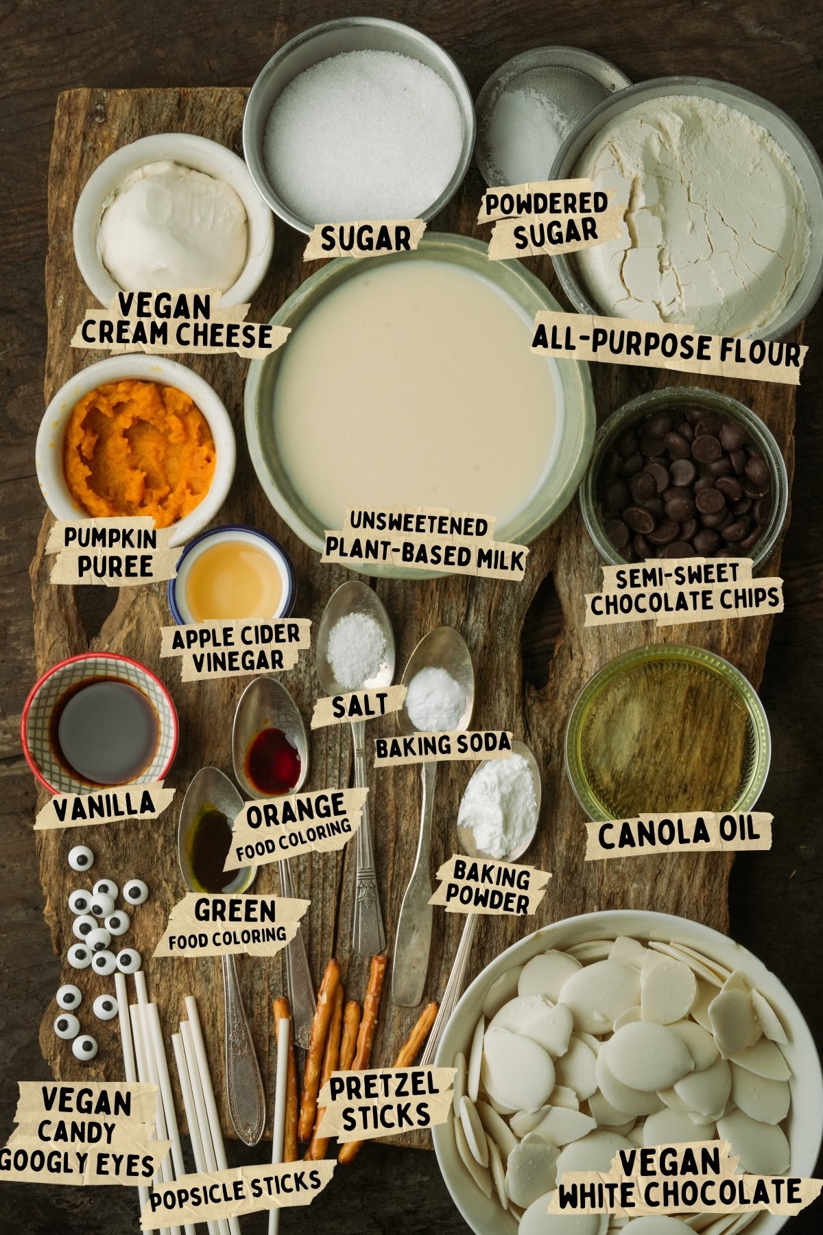 Wooden board with labeled ingredients for making vegan Halloween cake pops including vegan cream cheese, pumpkin puree, sugars, flour, chocolate chips, plant-based milk, canola oil, food coloring, pretzel sticks, and candy googly eyes.