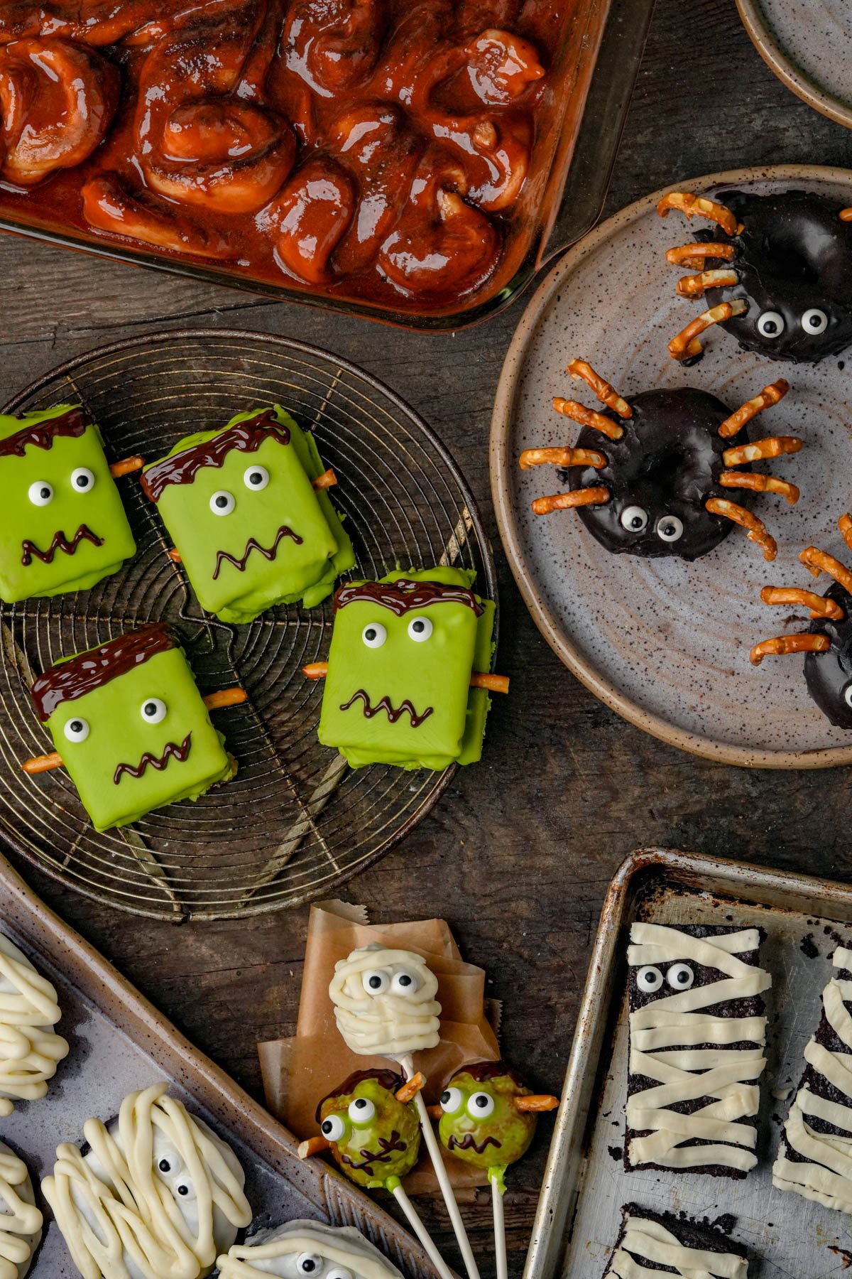 Assorted Halloween-themed treats including green Frankenstein s'mores, spider donuts with pretzel legs, mummy cake pops, and a dish of cinnamon roll guts.