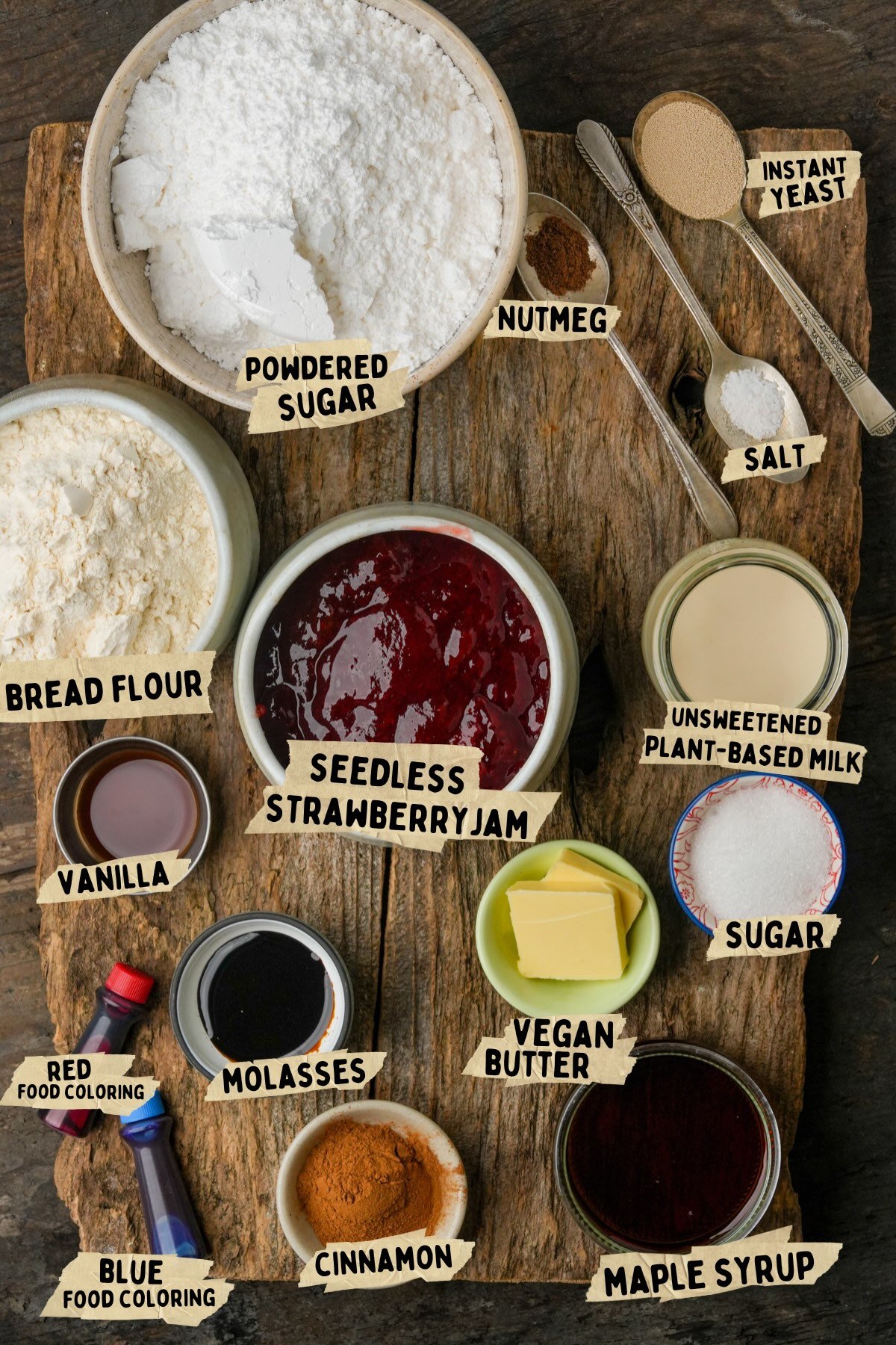 Various ingredients for a vegan Halloween cinnamon rolls recipe including powdered sugar, seedless strawberry jam, bread flour, instant yeast, spices, and syrups, are arranged on a wooden board and labeled.