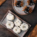 Plate of spider-shaped vegan Halloween donuts with pretzel legs and chocolate coating, and plate of mummy-shaped donuts with white icing and candy eyes on a wooden table.
