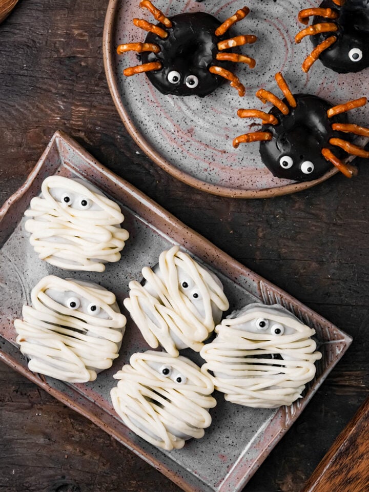 Plate of spider-shaped vegan Halloween donuts with pretzel legs and chocolate coating, and plate of mummy-shaped donuts with white icing and candy eyes on a wooden table.