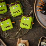 Vegan Halloween s'mores decorated as green Frankenstein faces with candy eyes and pretzel stick bolts are arranged on a cooling rack and plate, alongside other Halloween-themed treats.