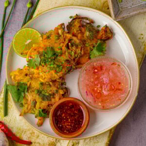 Three vegetable okoy fritters on a white plate, garnished with chopped herbs, served with a lime wedge, a bowl of chili vinegar, and a bowl of red chili sauce.
