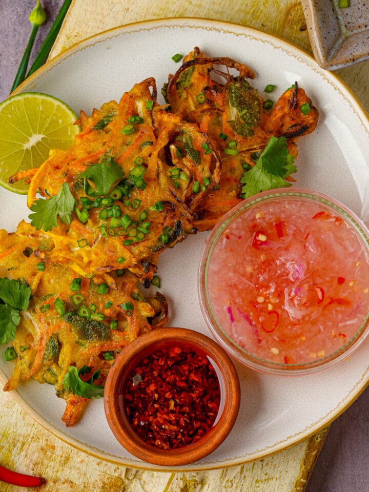 Three vegetable okoy fritters on a white plate, garnished with chopped herbs, served with a lime wedge, a bowl of chili vinegar, and a bowl of red chili sauce.