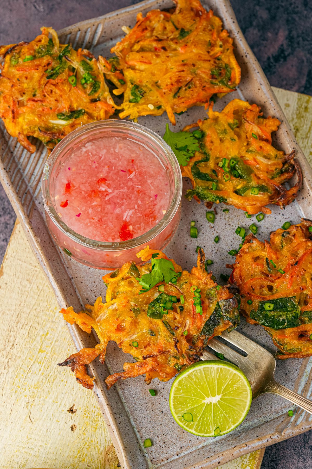 A plate of vegetable okoy fritters served with a small jar of pink dipping sauce, garnished with chopped herbs, and a halved lime on the side.