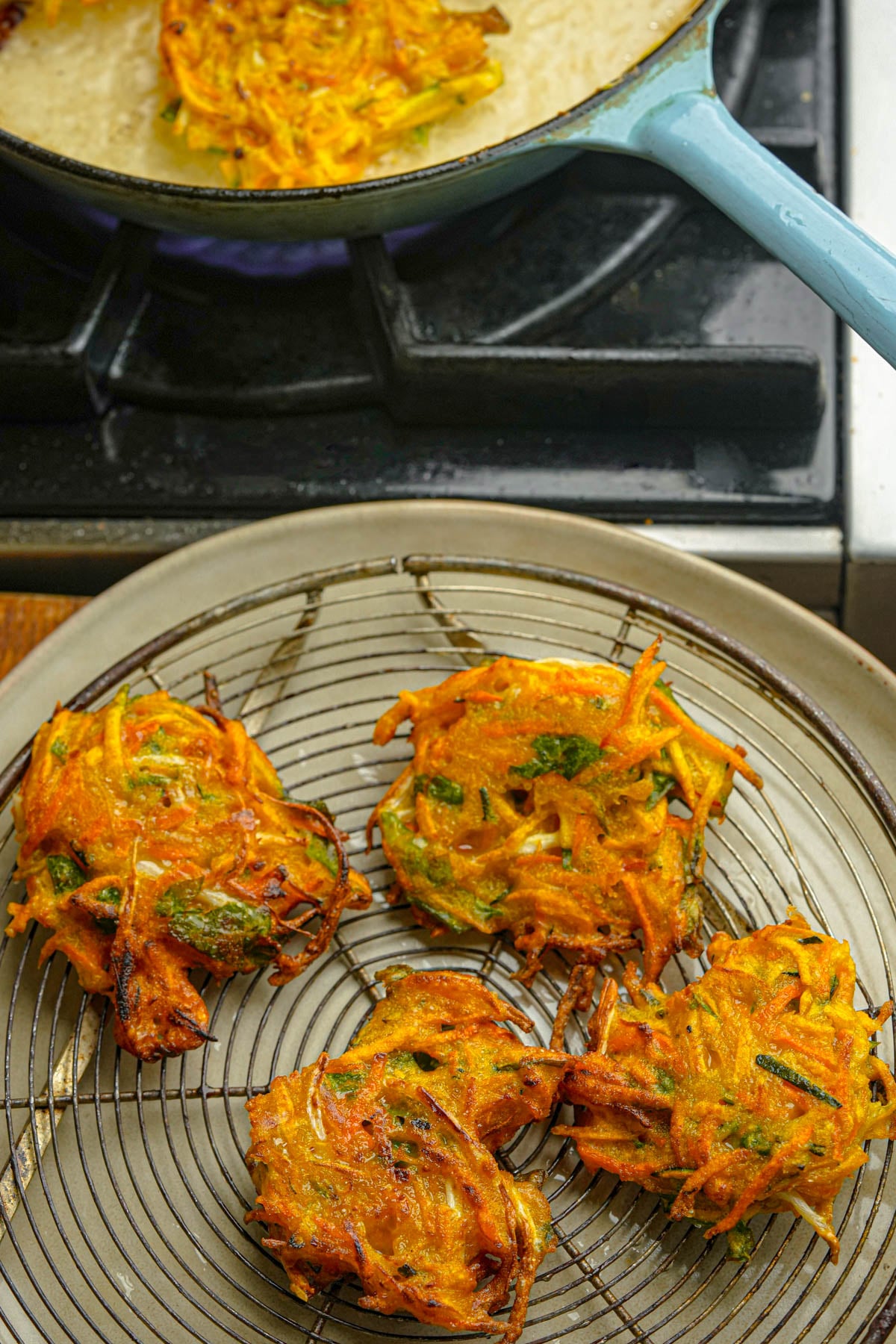 Four vegetable okoy fritters are cooling on a wire rack over a tray, with a pan of oil and another fritter visible in the background on the stove.