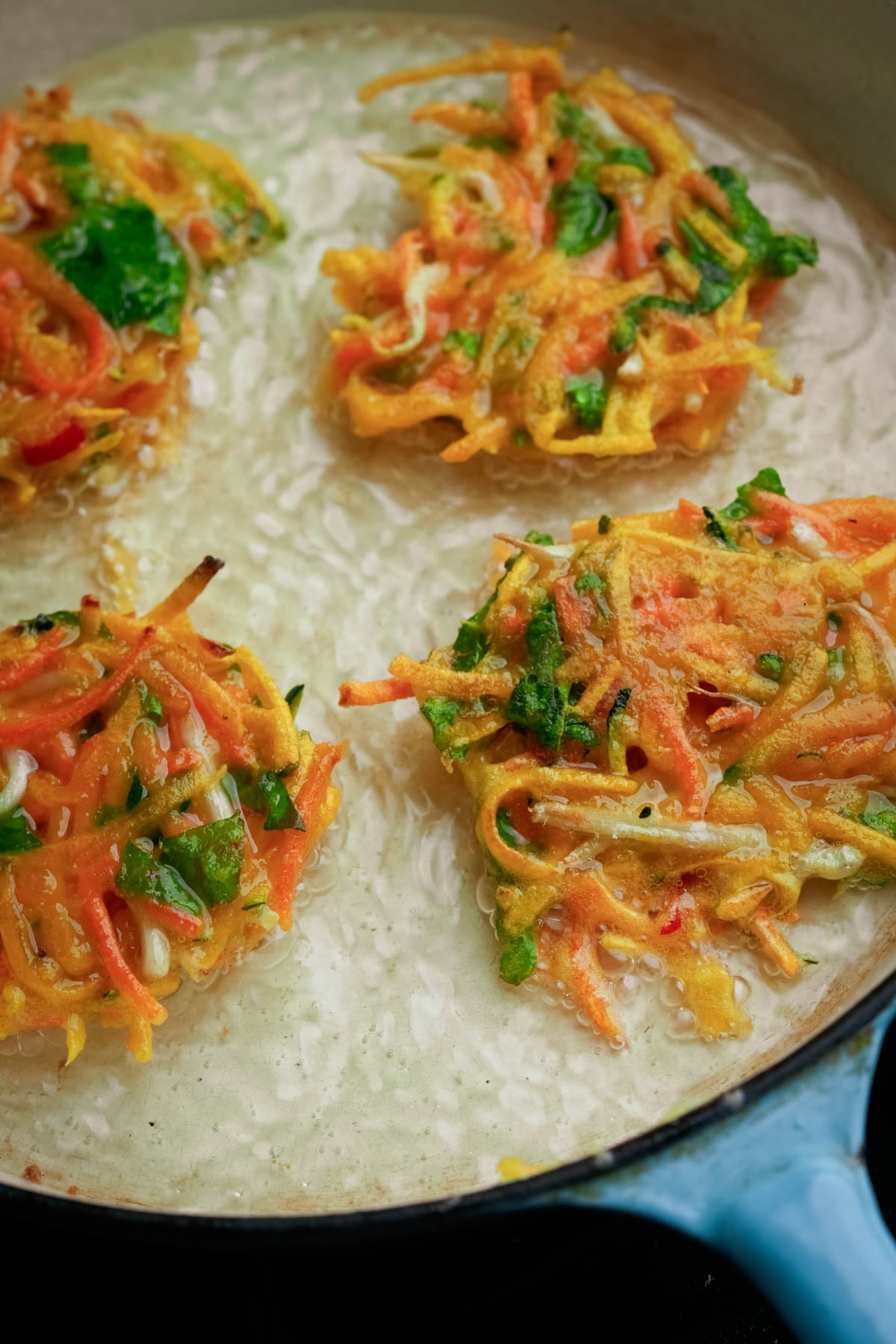 Four vegetable okoy fritters with shredded carrots and greens are frying in oil in a pan.
