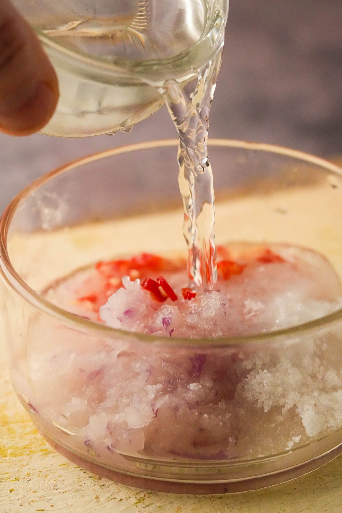 Clear liquid being poured from a small glass bowl into a larger bowl containing shredded red onion and chopped red chili peppers to make a pink chili vinegar to dip vegetable okoy into.