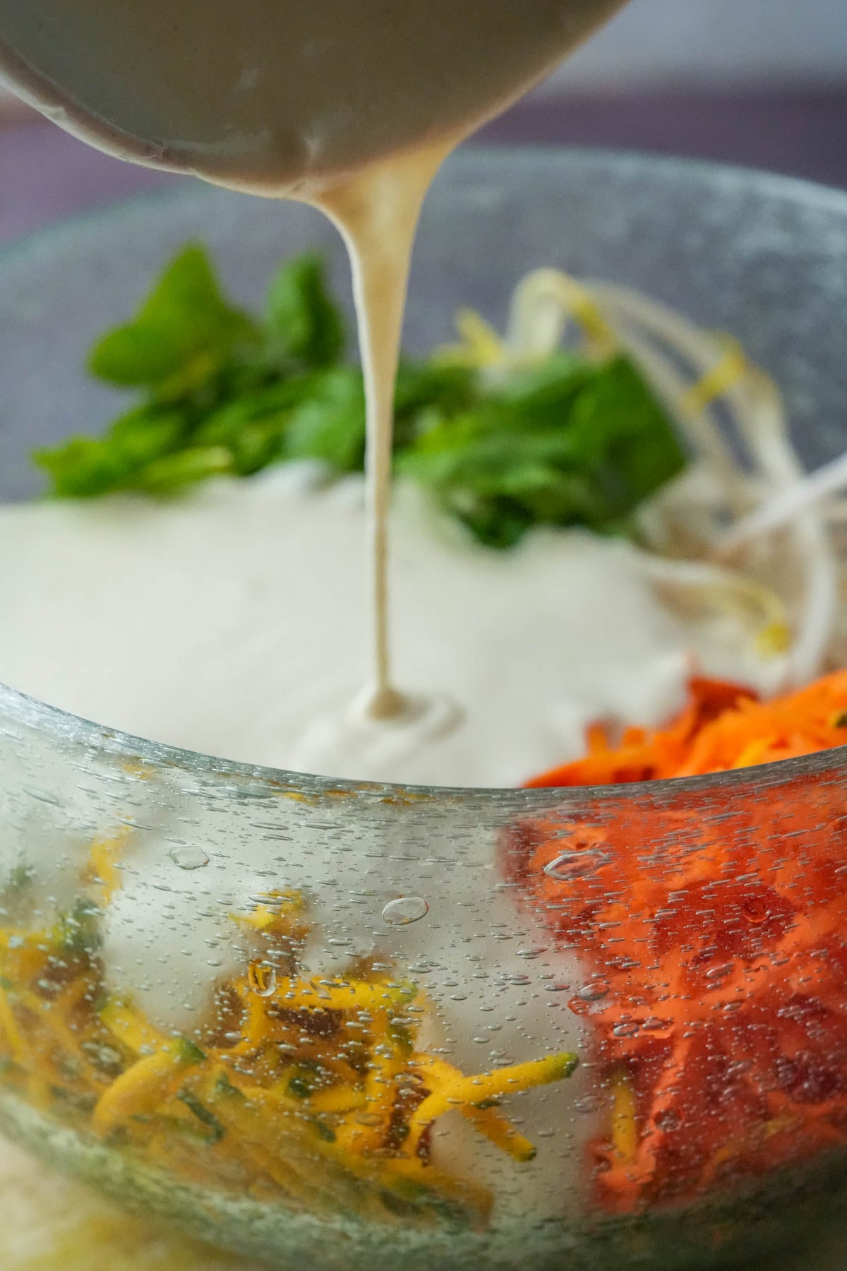A bowl of mixed batter is being poured into a glass bowl containing shredded carrots, kabocha, bean sprouts, and chopped spinach to make vegetable okoy with.