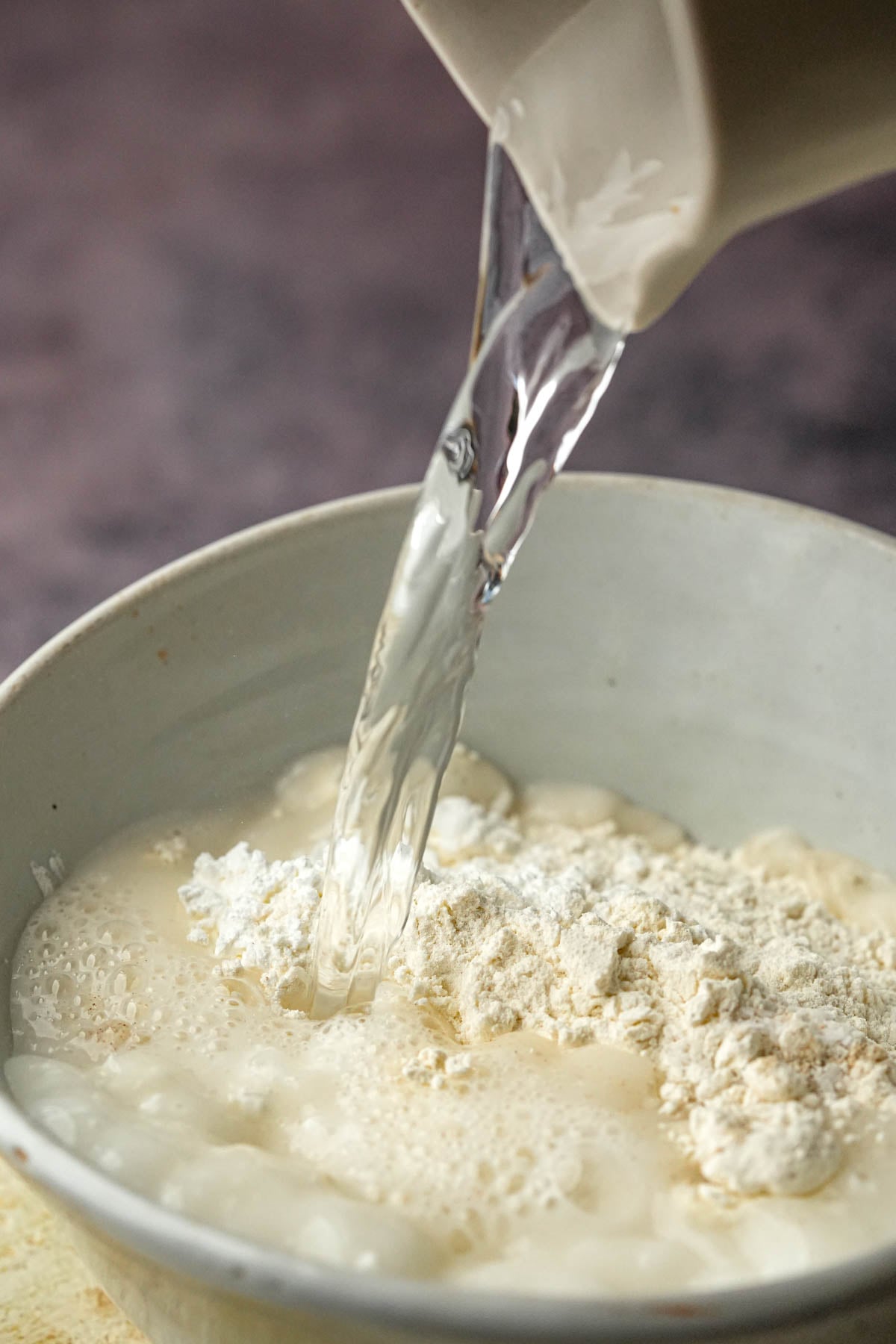 A stream of water is being poured from a jug into a bowl containing flour and other dry ingredients to make a vegetable okoy batter.