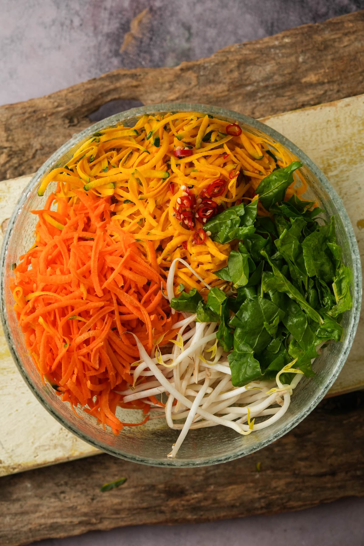 A glass bowl contains shredded carrot, kabocha squash, chopped spinach, bean sprouts, and sliced red chili, arranged in separate sections.