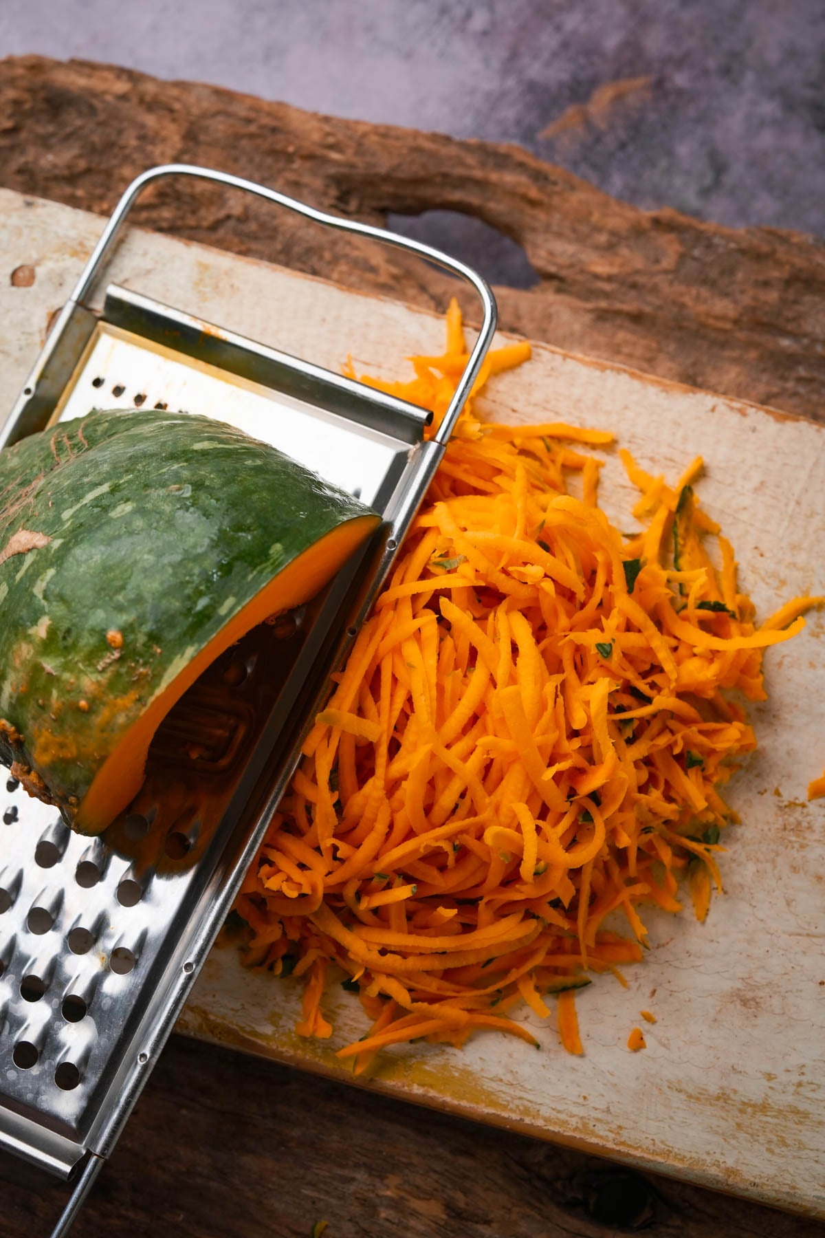 A chunk of kalabasa being grated on a metal grater, with a pile of freshly shredded pumpkin on a wooden cutting board.