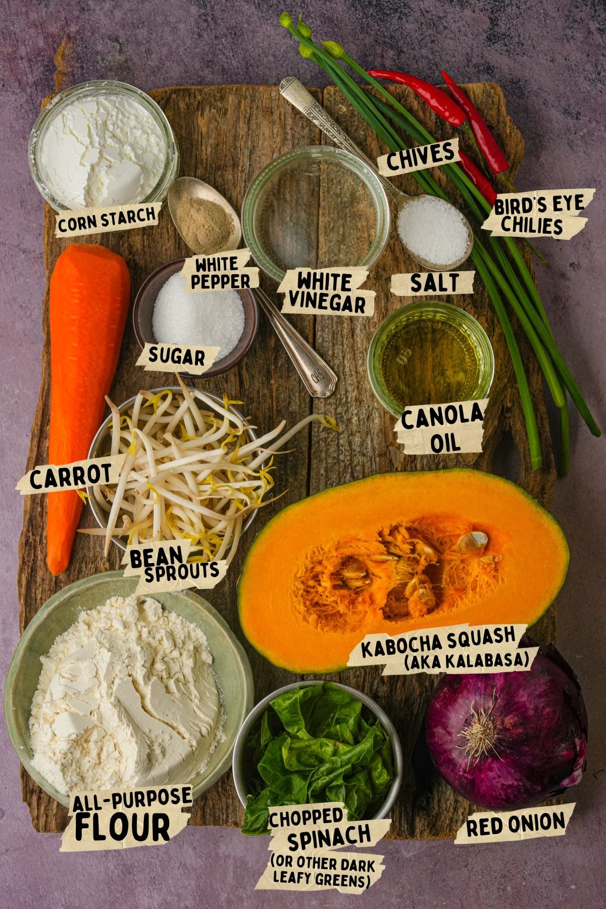 A variety of labeled ingredients for a vegetable okoy recipe on a wooden surface, including kabocha squash, carrot, red onion, flour, bean sprouts, spinach, chives, chilies, white vinegar, white pepper, salt, and canola oil.