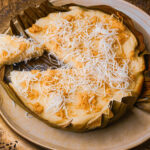 A round Filipino bibingka cake, topped with grated coconut and golden brown melted cheese, served on a plate and wrapped in banana leaves. A slice has been cut and slightly separated from the cake.