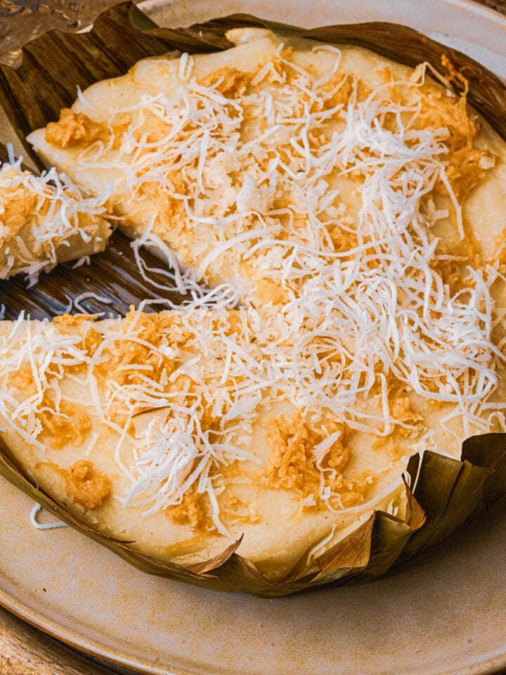 A round Filipino bibingka cake, topped with grated coconut and golden brown melted cheese, served on a plate and wrapped in banana leaves. A slice has been cut and slightly separated from the cake.