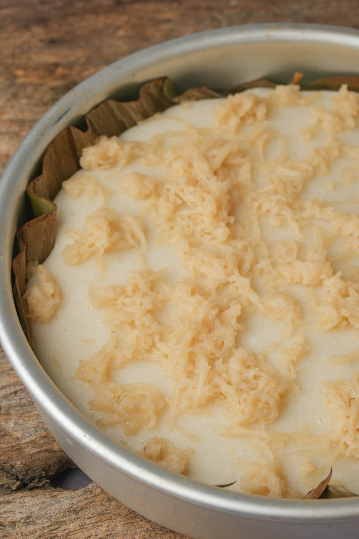 A round metal baking pan with a steamed Filipino bibingka cake, topped with shredded vegan cheese and set on a rustic wooden surface.