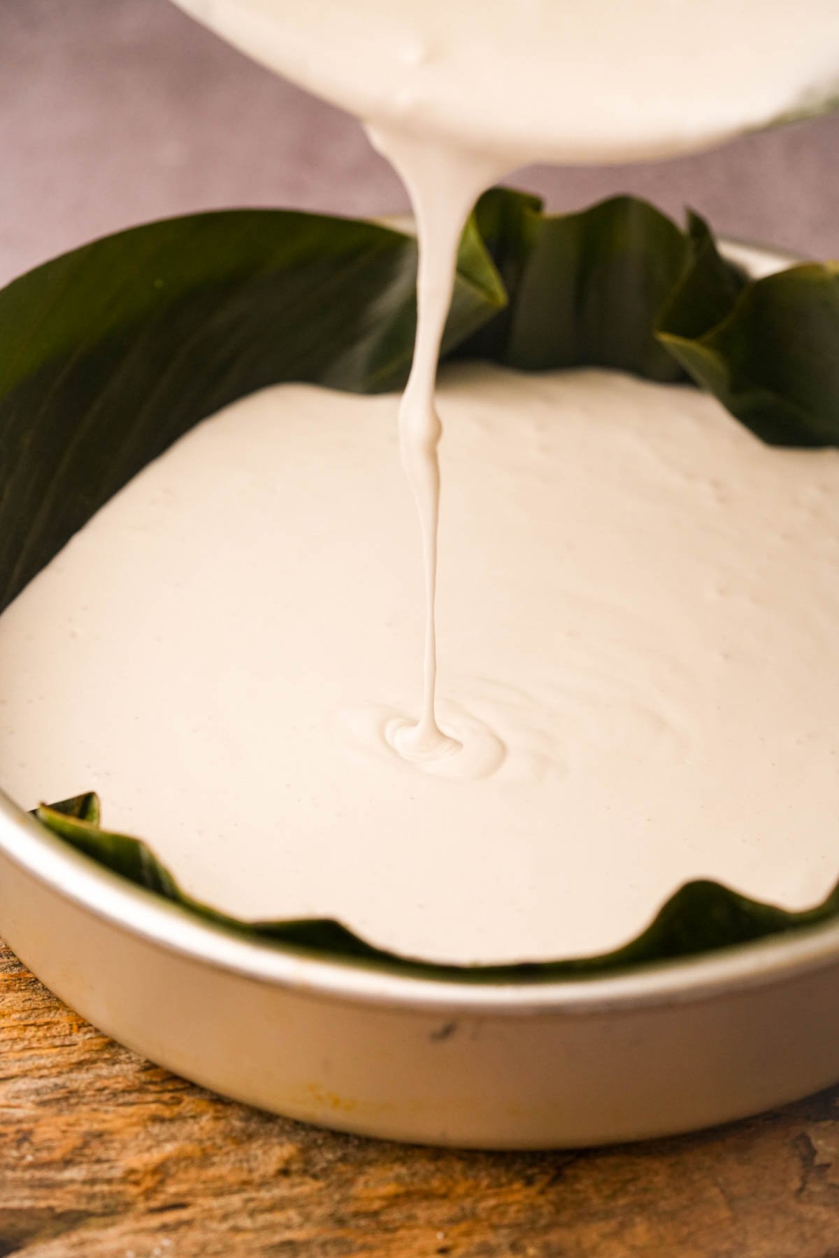 Thick white bibingka batter is being poured into a round baking pan lined with banana leaves, on a wooden surface.