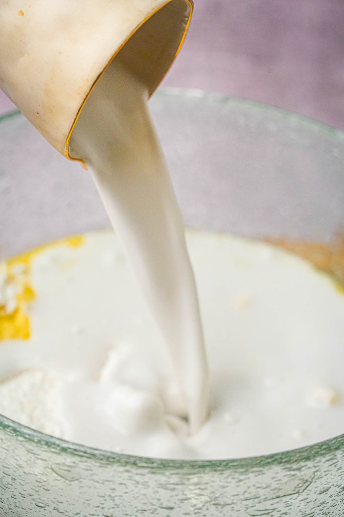 A close-up of coconut milk being poured from a cup into a glass bowl containing other ingredients.