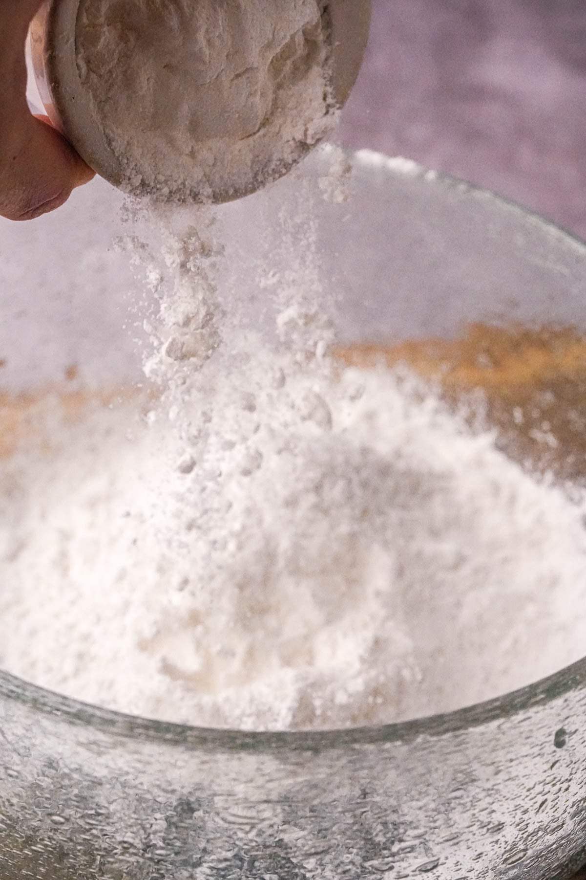 A hand pours rice flour from a measuring cup into a clear glass mixing bowl filled with more flour, with some flour particles falling in mid-air.