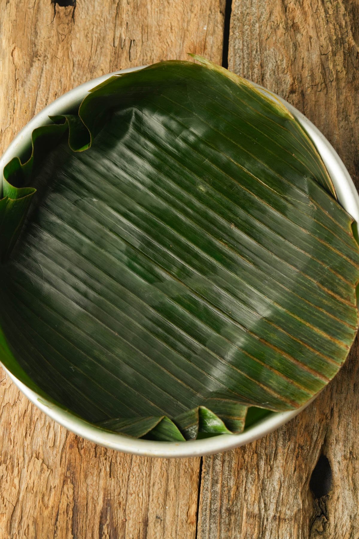 A white bowl lined with a large green banana leaf sits on a rustic wooden surface. The leaf covers the inside of the bowl, with its edges folded along the rim.