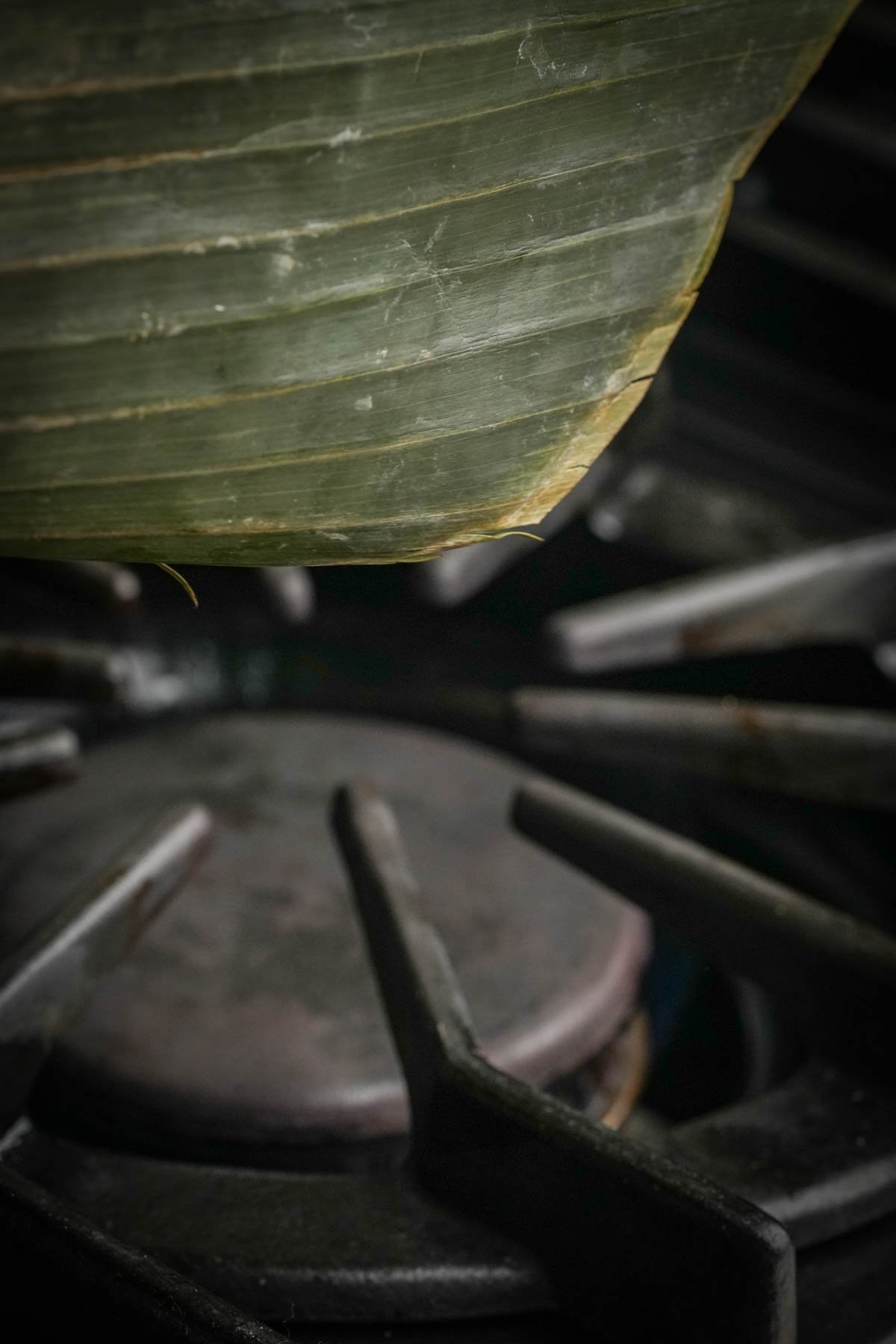 A close-up of a green banana leaf partially covering a dark, metal gas stove burner with visible grates. The image highlights the textures of both the leaf and the stove.